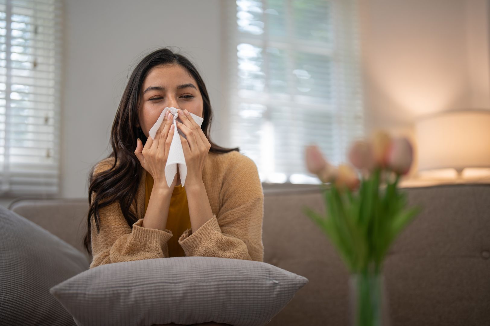 Woman sneezing into tissue sitting on couch, tulips on table